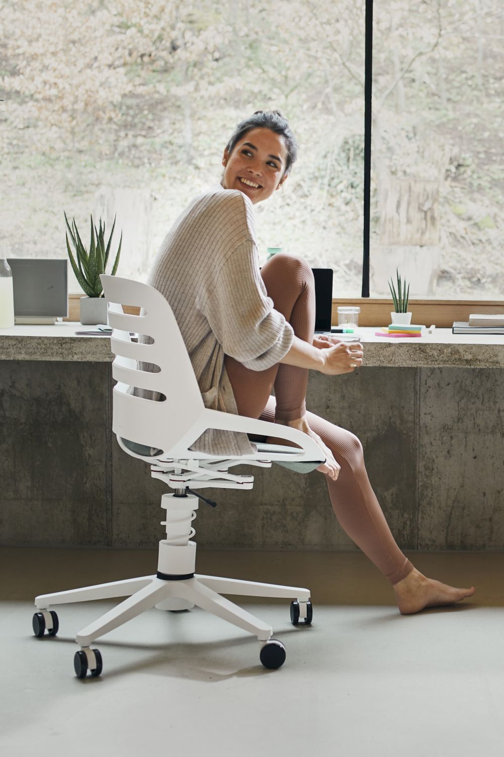 Frau sitzt lächelnd auf ergonomischem Bürostuhl vor einem großen Fenster, natürliches Licht im Hintergrund.