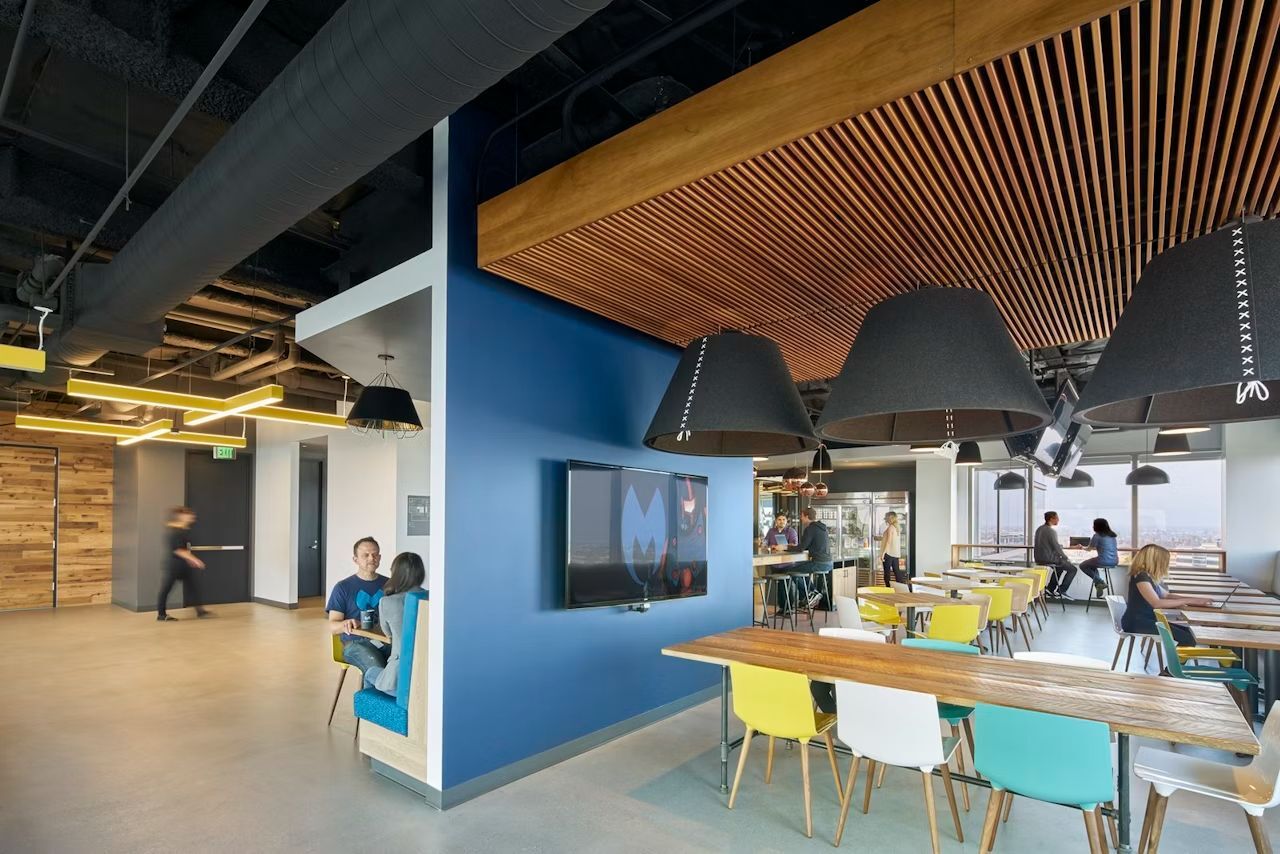 Moderner Bürokantinenbereich mit bunten Stühlen, großen Lampen und Personen, die an Tischen sitzen und reden.