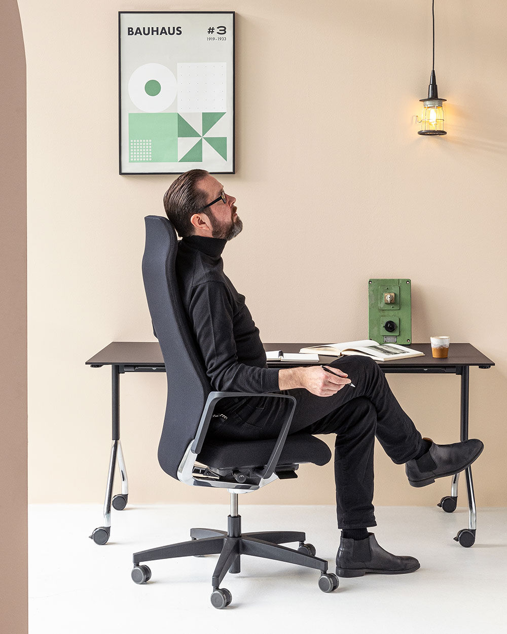 Mann sitzt entspannt in einem ergonomischen Bürostuhl vor einem Schreibtisch mit Bauhaus-Poster an der Wand.