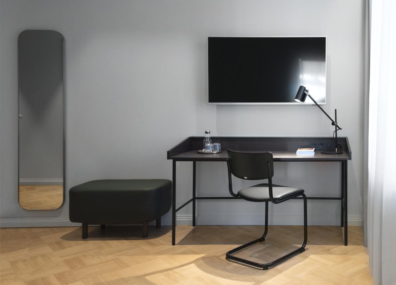 Modernes Büro mit ergonomischem Stuhl, Tischlampe, Spiegel, Hocker und großem Bildschirm an der Wand.