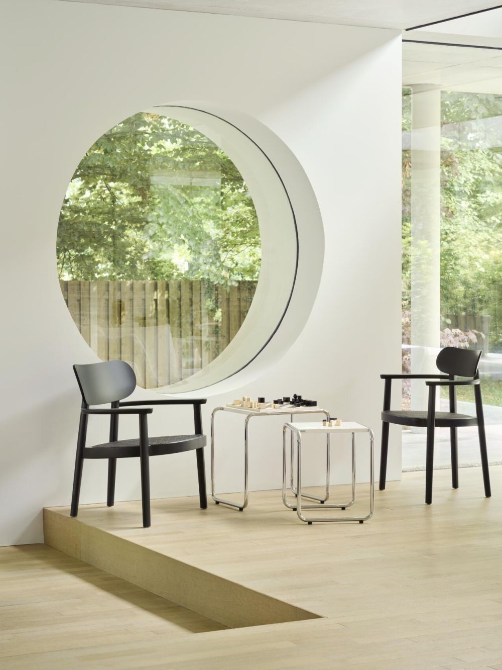 Zwei moderne Stühle und ein Tisch mit Schachspiel vor einem großen runden Fenster mit Gartenblick.