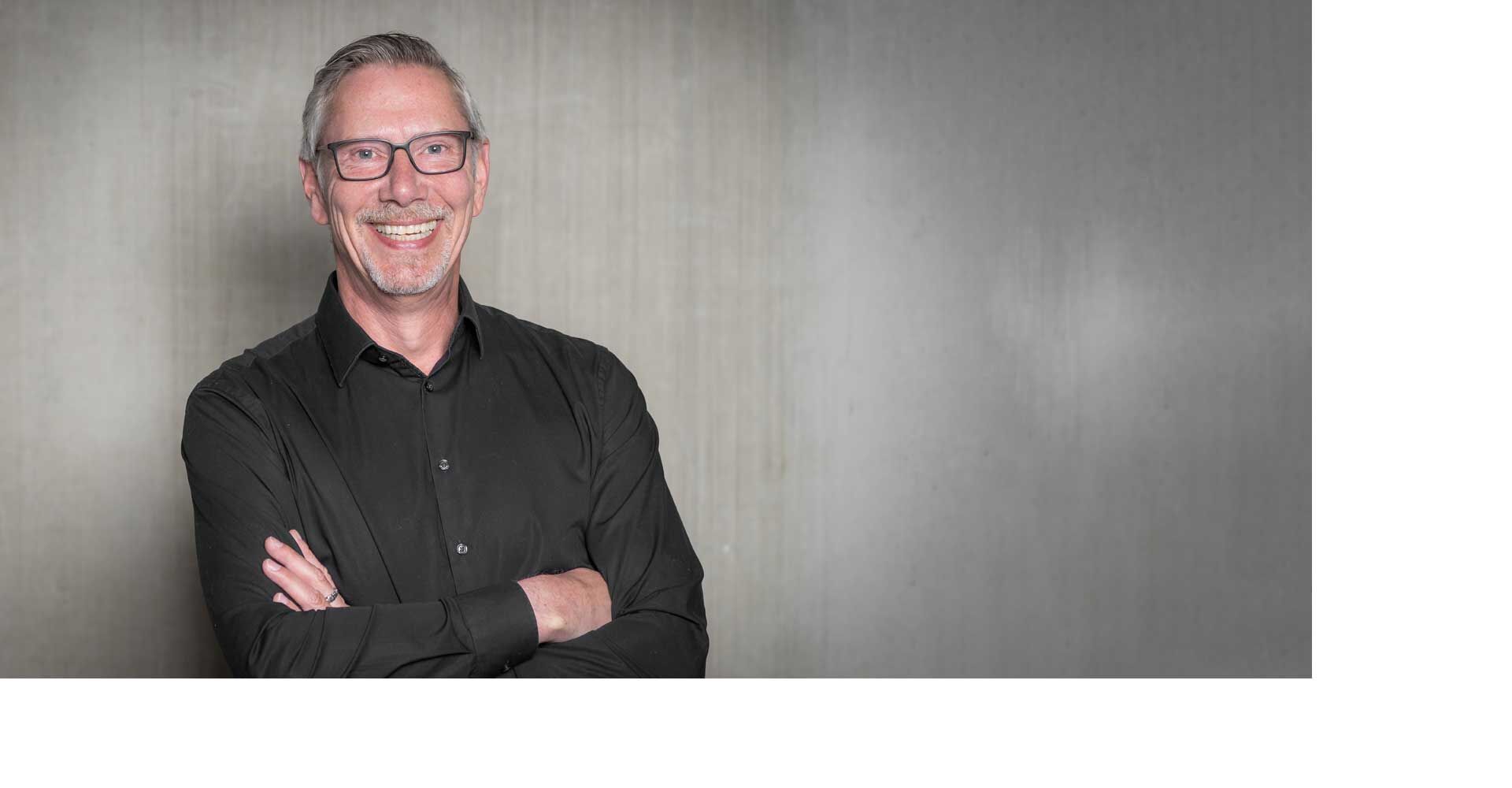 Mann mit Brille lächelt, trägt ein schwarzes Hemd, steht vor einer grauen Wand.