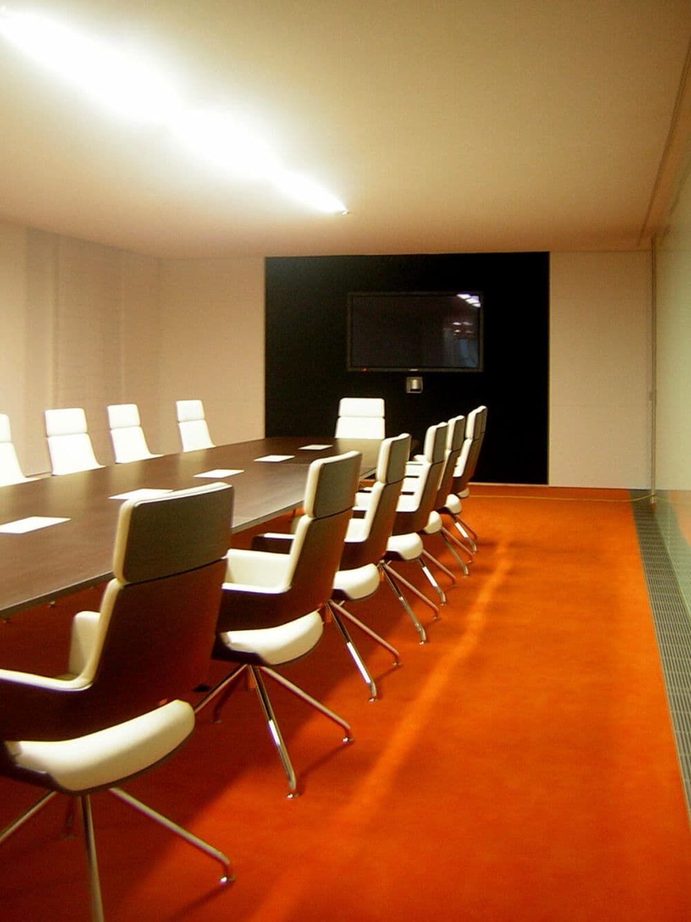 Moderner Konferenzraum mit ergonomischen Bürostühlen um einen langen Holztisch, orangefarbener Teppich und Wandmonitor.