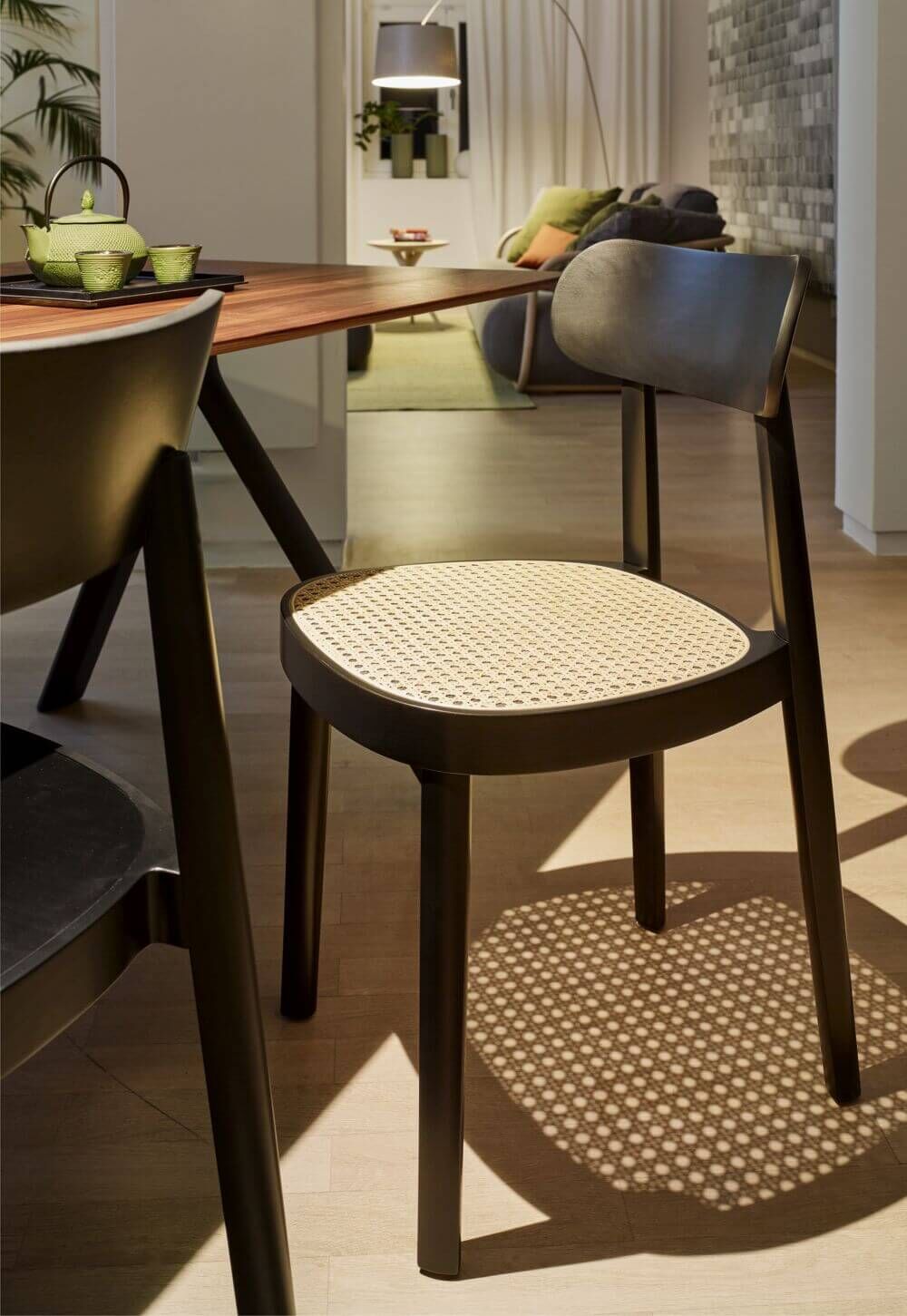 Moderner Holzstuhl mit geflochtener Sitzfläche in gemütlichem Wohnzimmer neben großem Holztisch, stimmungsvolles Licht.