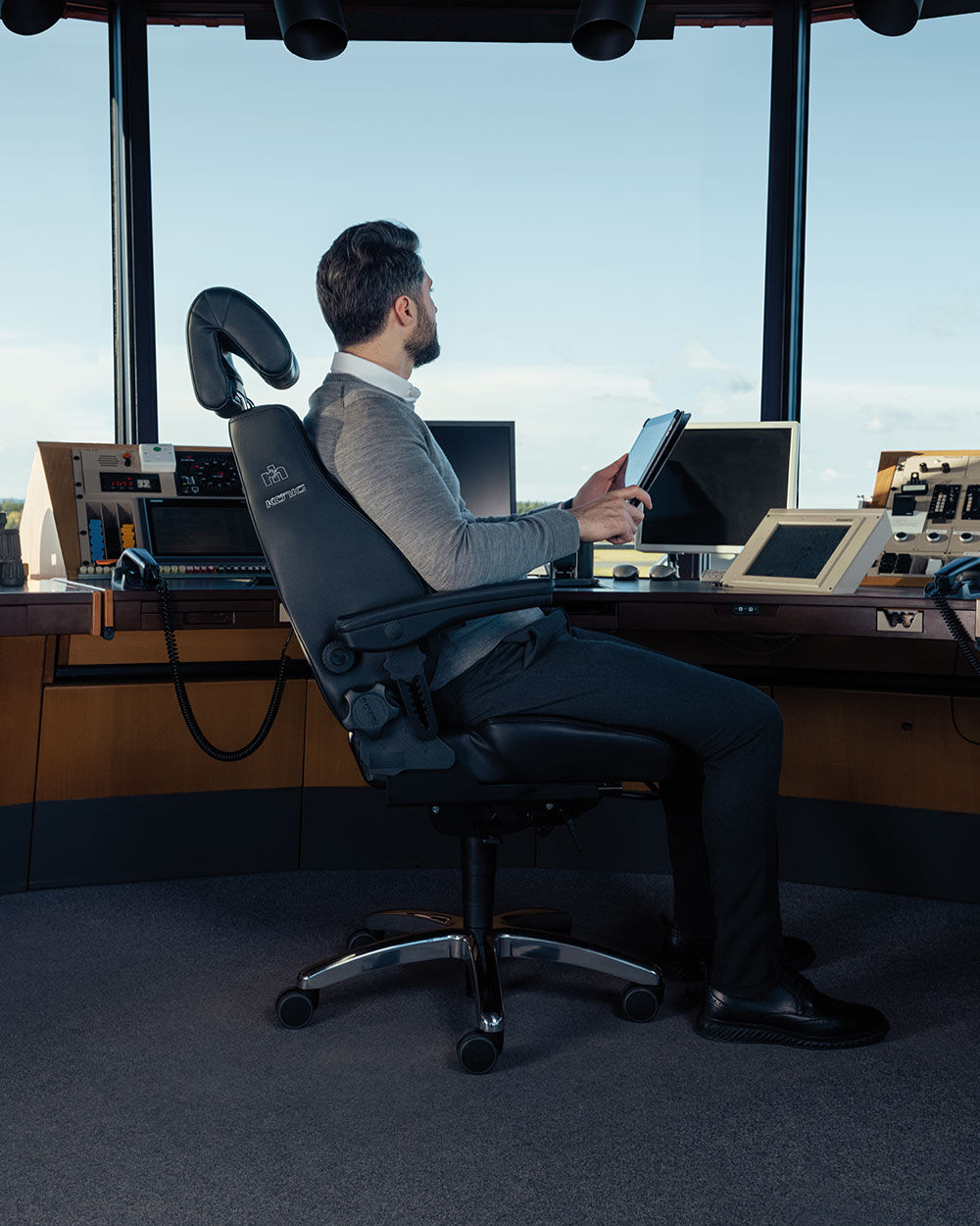 Mann sitzt ergonomisch auf Bürostuhl, hält Tablet, vor modernem Arbeitsplatz mit Computerbildschirmen und Fensterblick.
