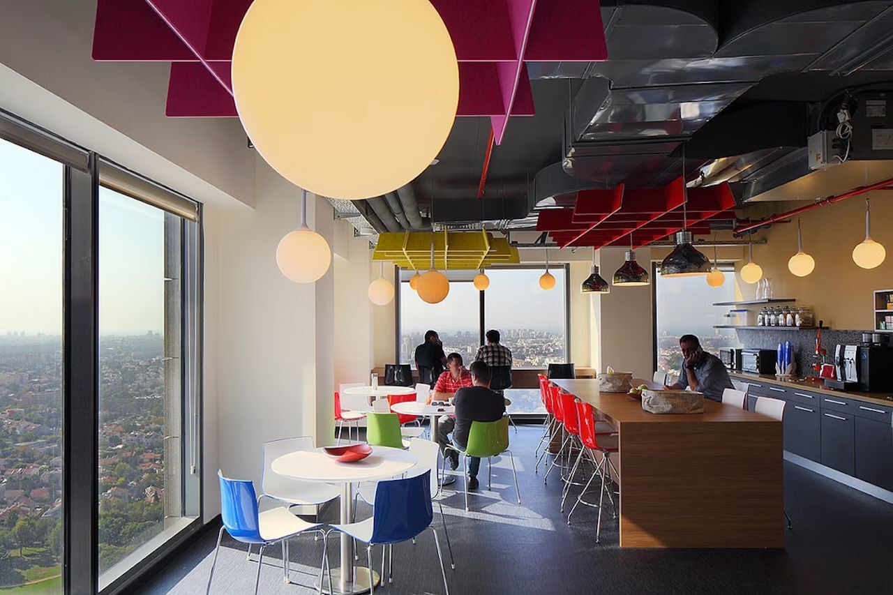 Moderne Büroküche mit farbigen Stühlen, großen Fenstern und kreativer Beleuchtung, Menschen sitzen und unterhalten sich.