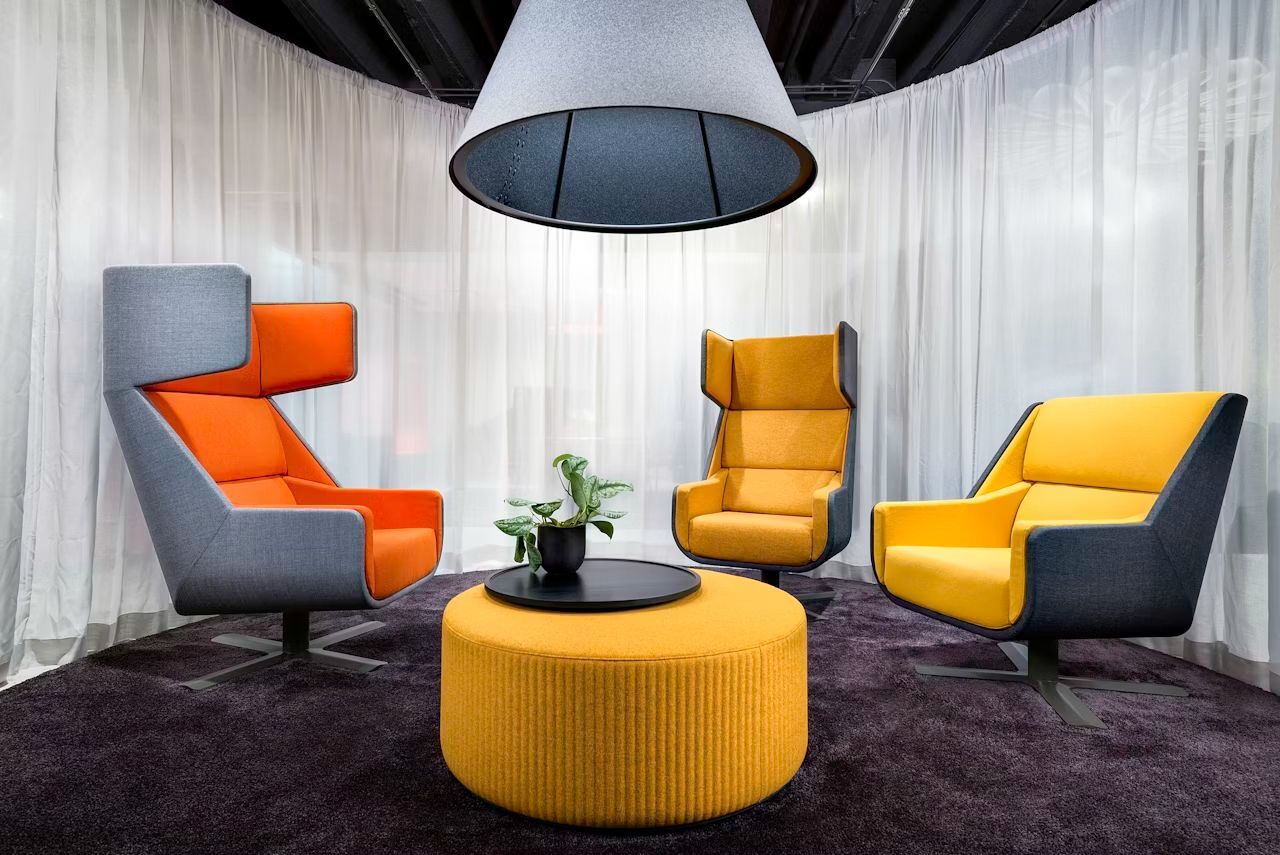 Drei ergonomische Sessel in Grau, Orange und Gelb um einen runden, gepolsterten Tisch mit Pflanze.