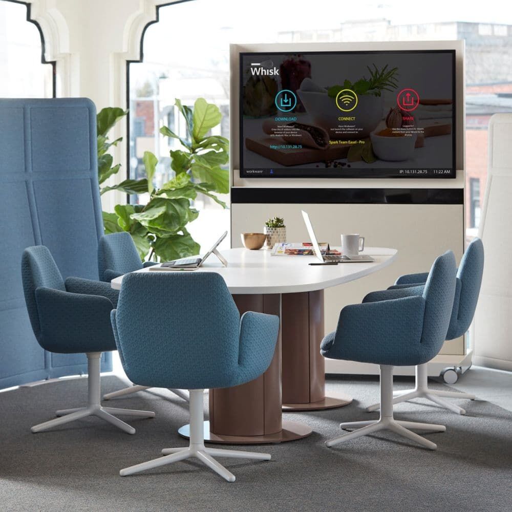 Moderner Besprechungsraum mit drehbaren blauen Stühlen, ovalem Tisch, großer Pflanze und Bildschirm im Hintergrund.
