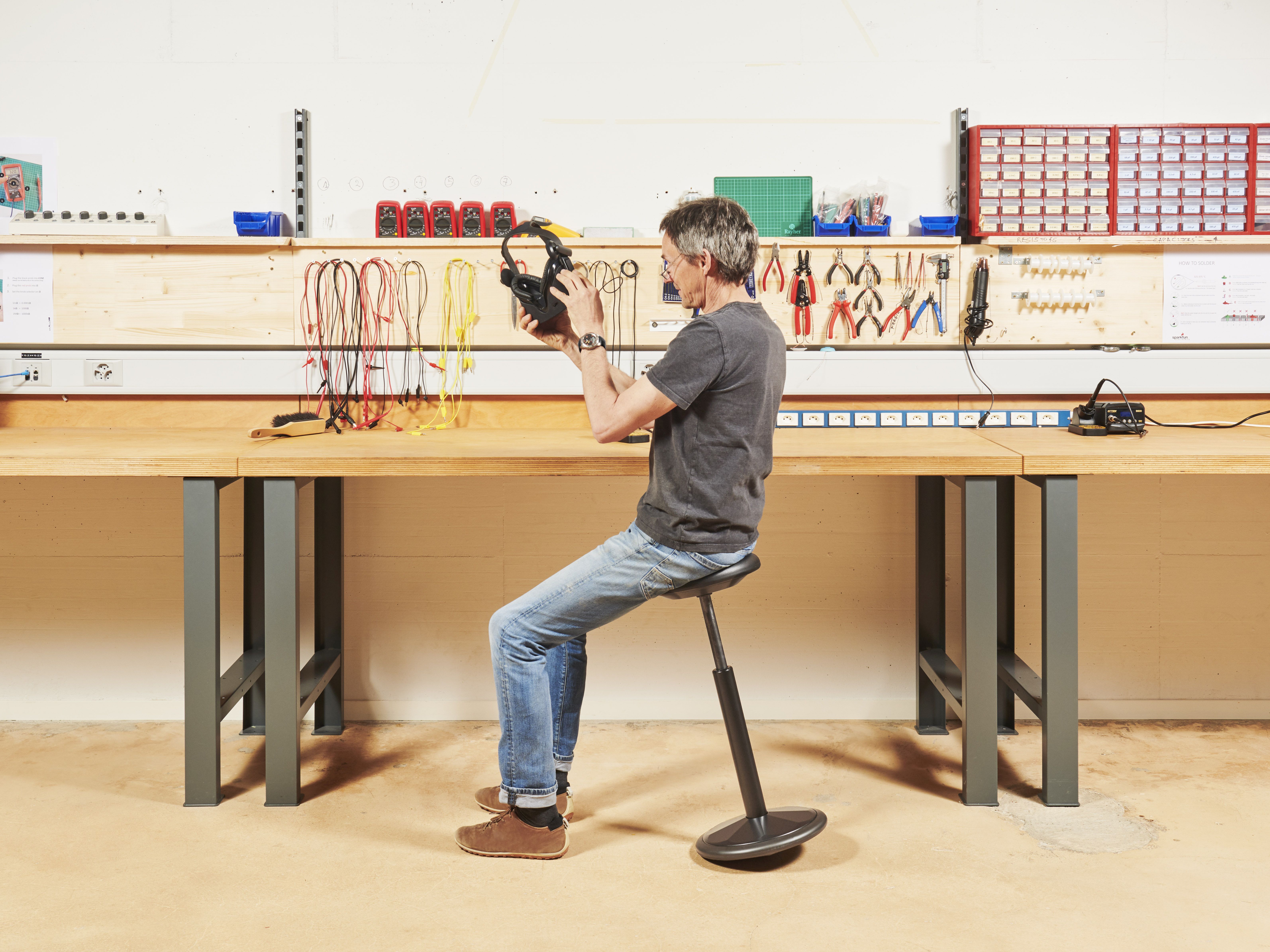 Person sitzt auf ergonomischem Hocker an einer Werkbank, umgeben von Werkzeugen und Kabeln.