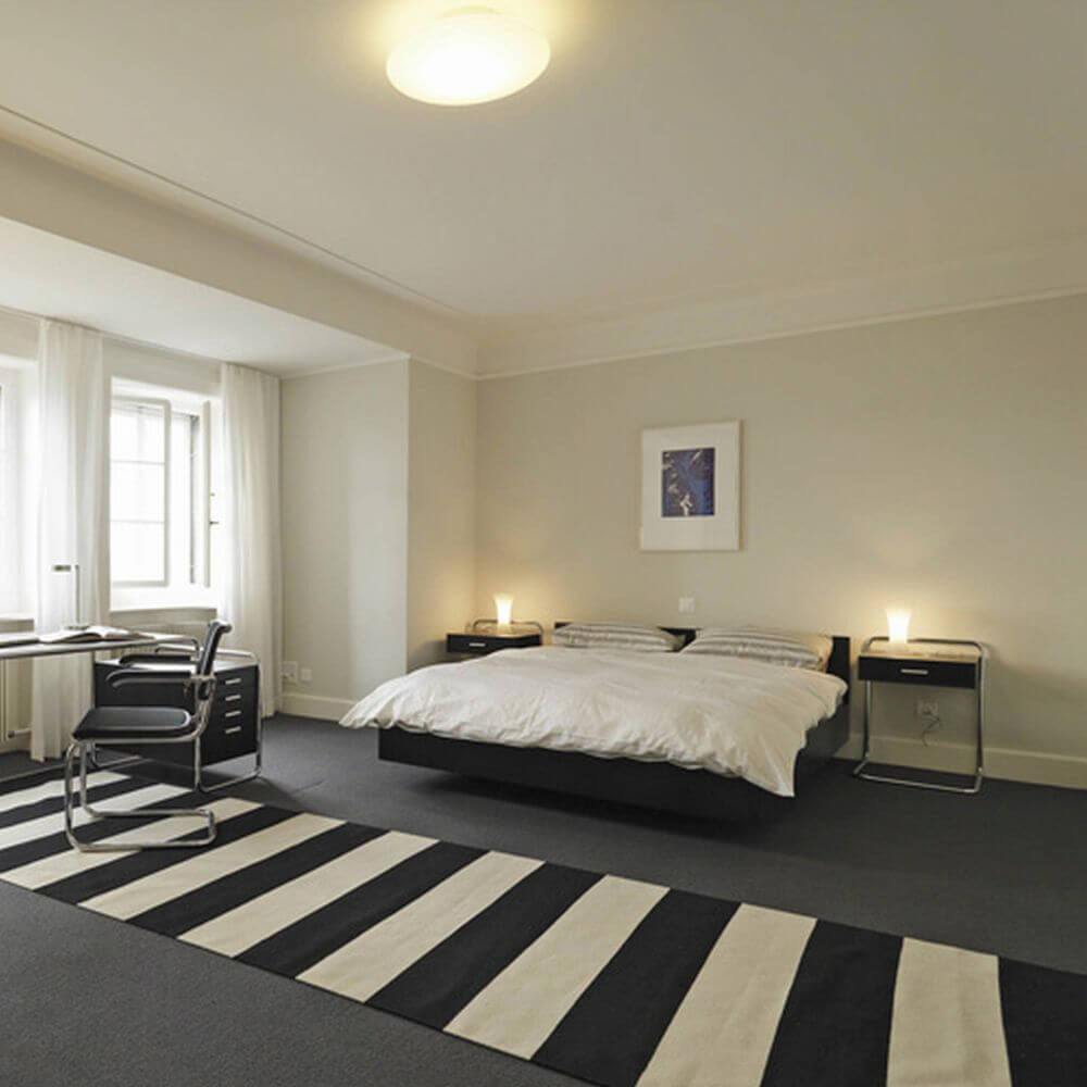 Modernes Schlafzimmer mit Bett, Schreibtisch und Streifenteppich, betont minimalistisches Design und Komfort.