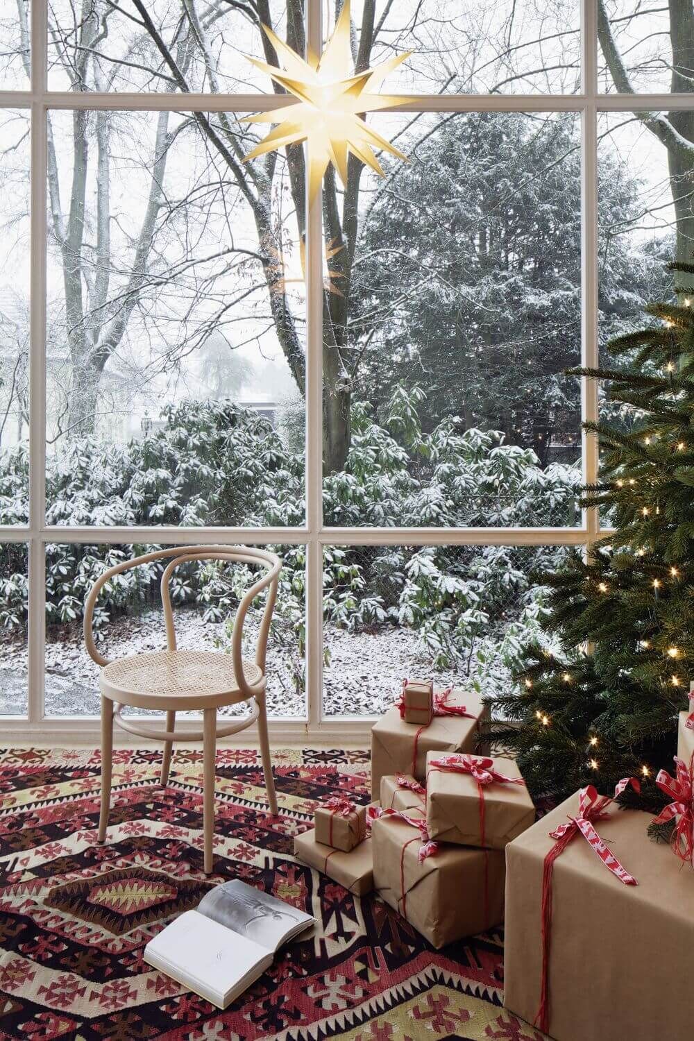 Ein ergonomischer Stuhl, Geschenke mit roten Schleifen und ein Weihnachtsbaum vor einem schneebedeckten Gartenfenster.