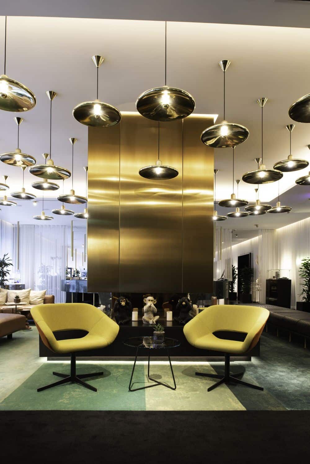 Modernes Wohnzimmer mit gelben Sesseln, Glastisch und eleganter Beleuchtung vor einem goldenen Metallelement.