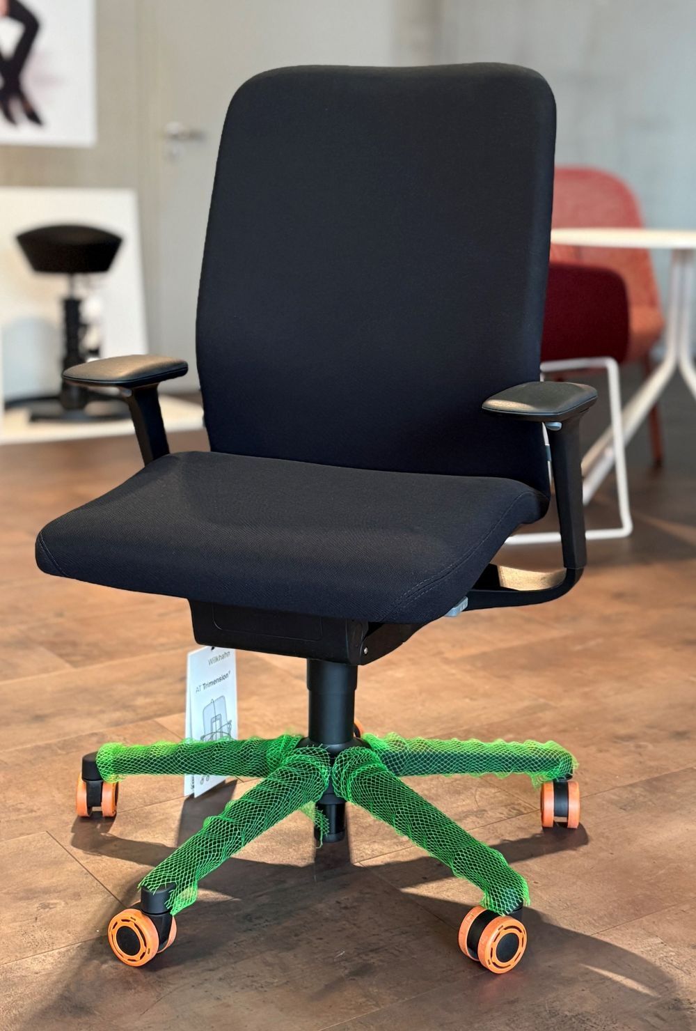 Ergonomischer Bürostuhl mit schwarzem Sitz und Armlehnen, grüne Abdeckung über der Drehbasis, vor modernem Hintergrund.