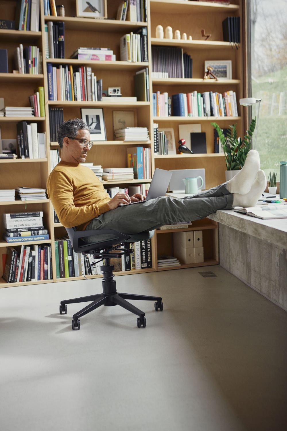 Mann in gelbem Pullover sitzt bequem auf ergonomischem Stuhl, Beine auf Tisch in Heimbüro mit Bücherregal.