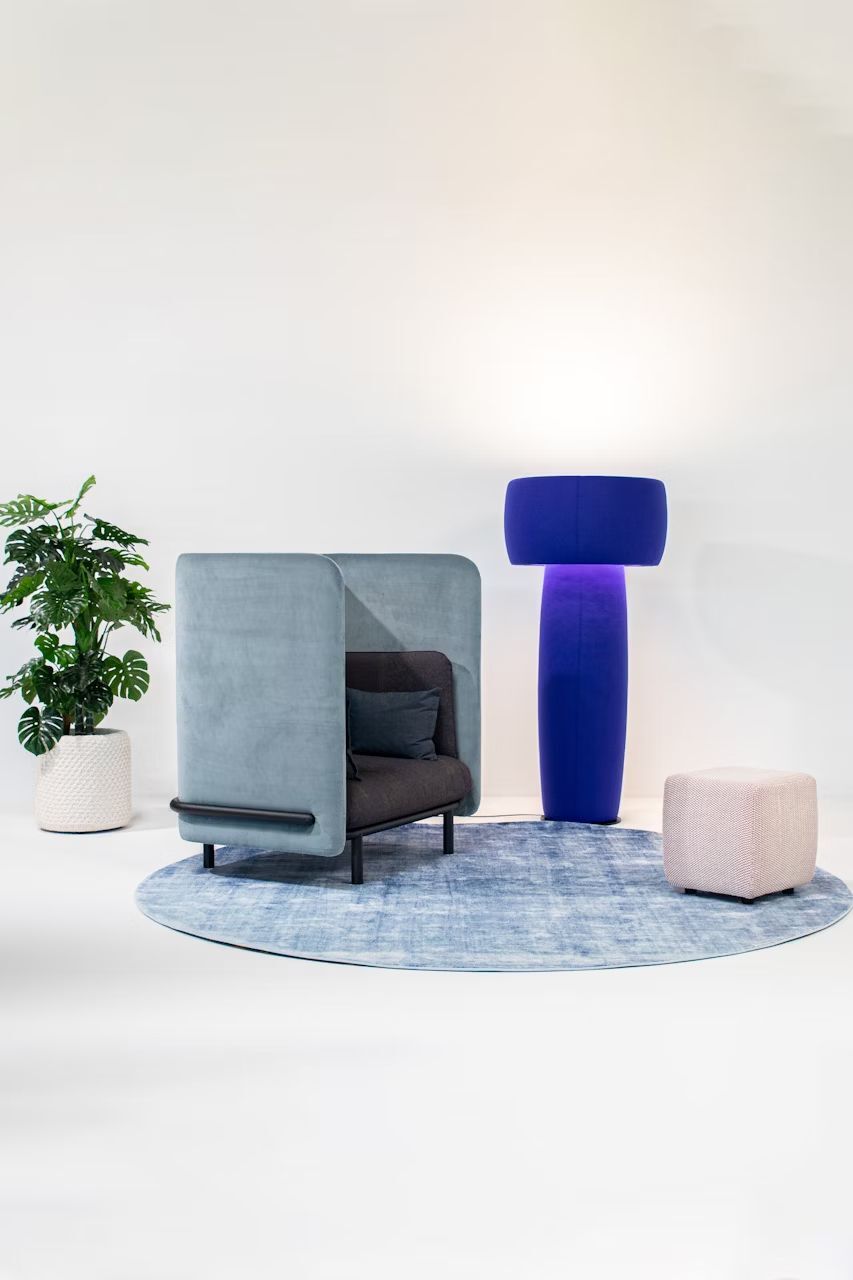 Ergonomischer Sessel mit hoher Rückenlehne, neben blauer Stehlampe und Pflanzen auf rundem Teppich.