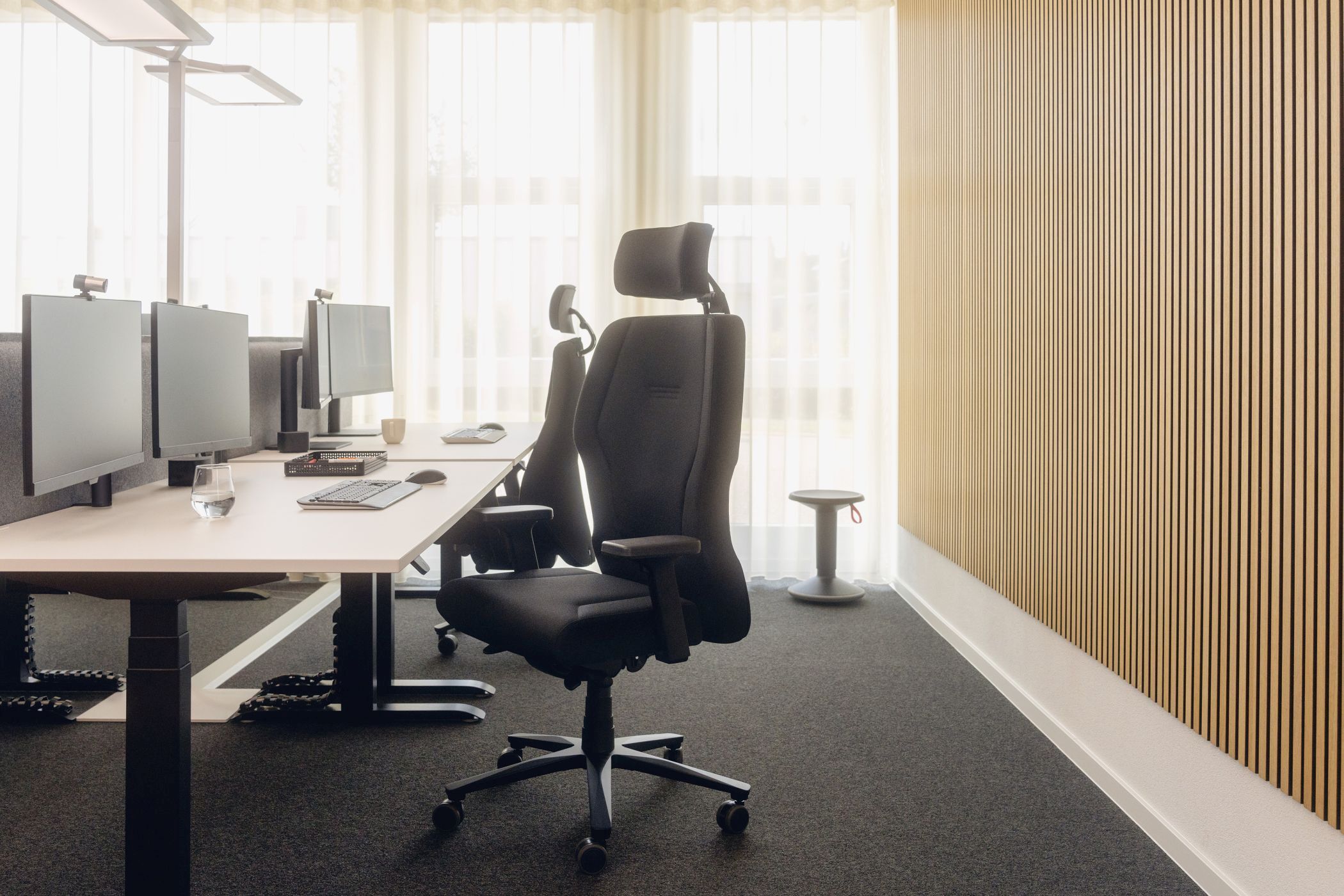 Moderner ergonomischer Bürostuhl in elegantem Büro mit mehreren Monitoren und natürlicher Beleuchtung.