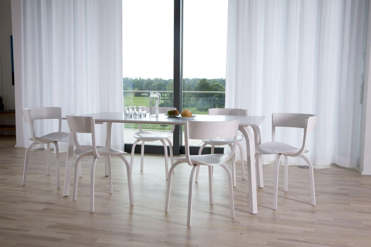 Moderner Essbereich mit weißem Tisch, sechs Stühlen und Blick auf eine grüne Landschaft durch Glasfenster.