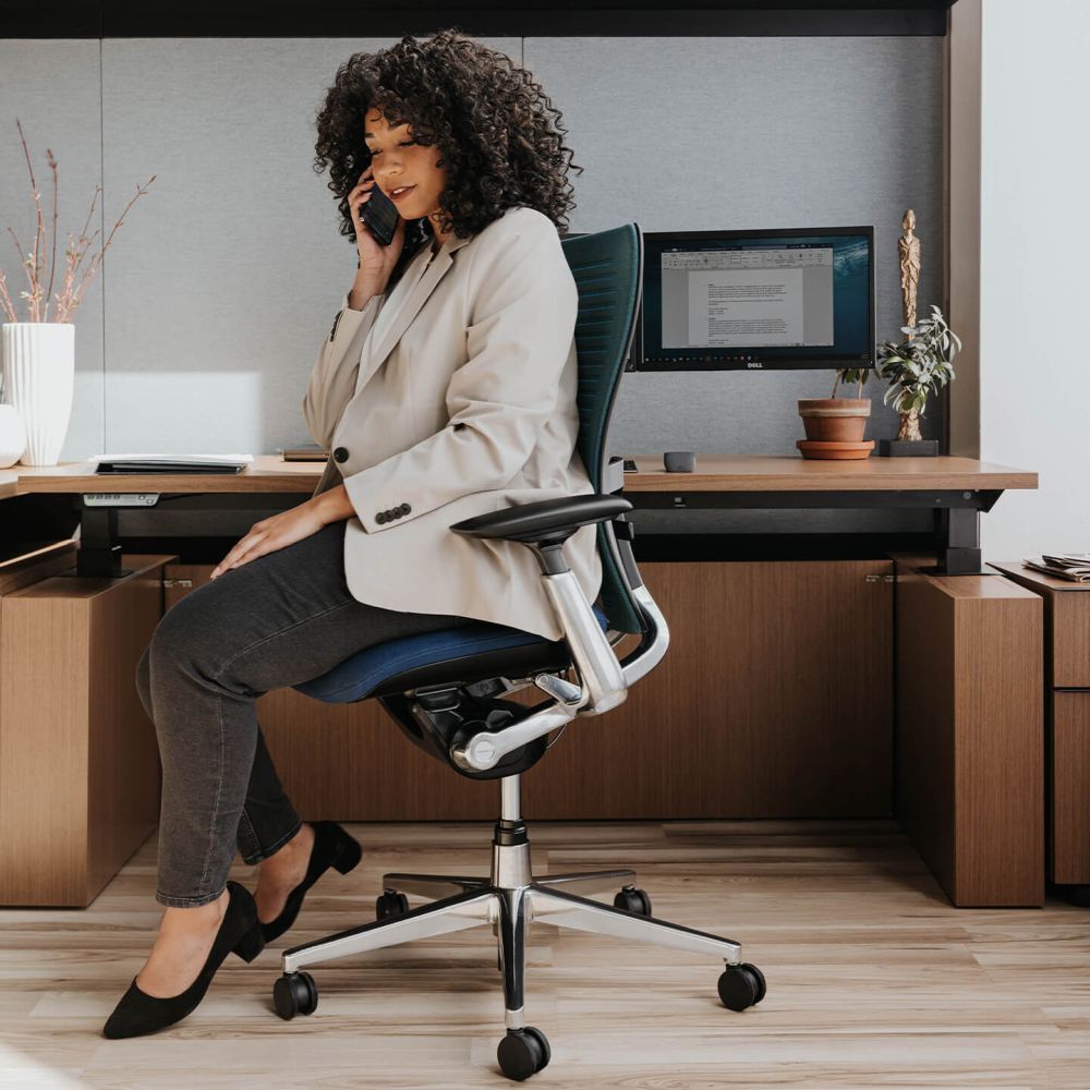 Frau sitzt auf ergonomischem Bürostuhl und telefoniert, Schreibtisch im Hintergrund, betont Produktqualität und Komfort.