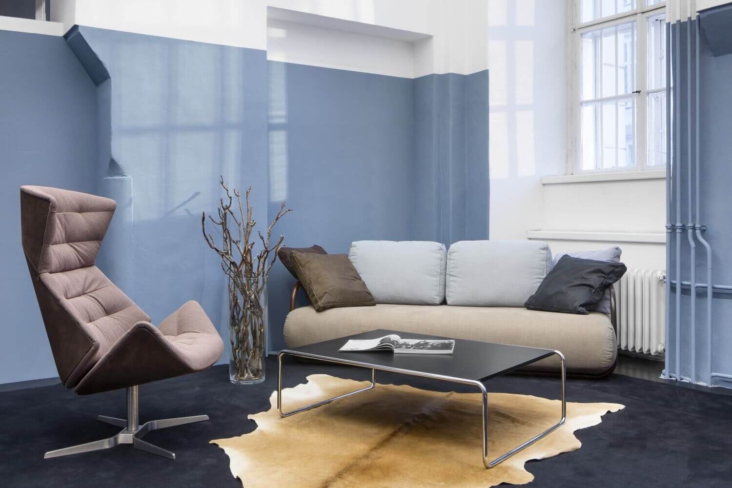 Modernes Wohnzimmer mit ergonomischem Stuhl, Sofa, Couchtisch und Teppich vor blauer Wand.