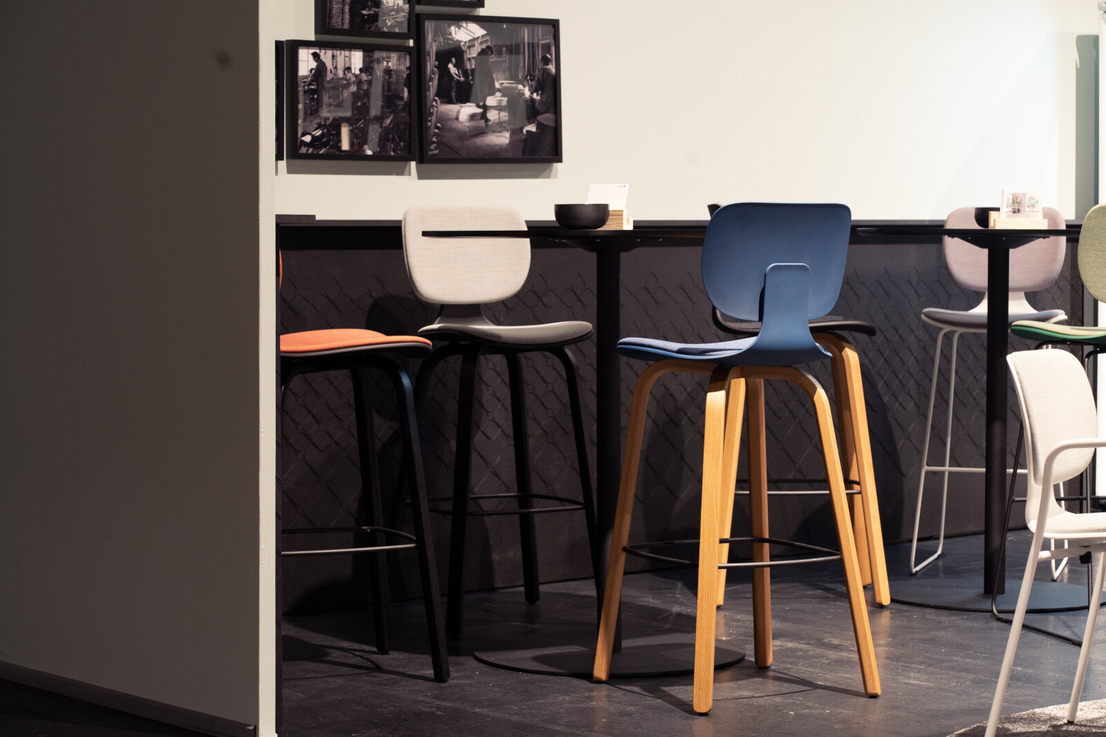Barhocker in verschiedenen Farben stehen an einem modernen Tisch, Betonwand und Schwarz-Weiß-Fotos im Hintergrund.