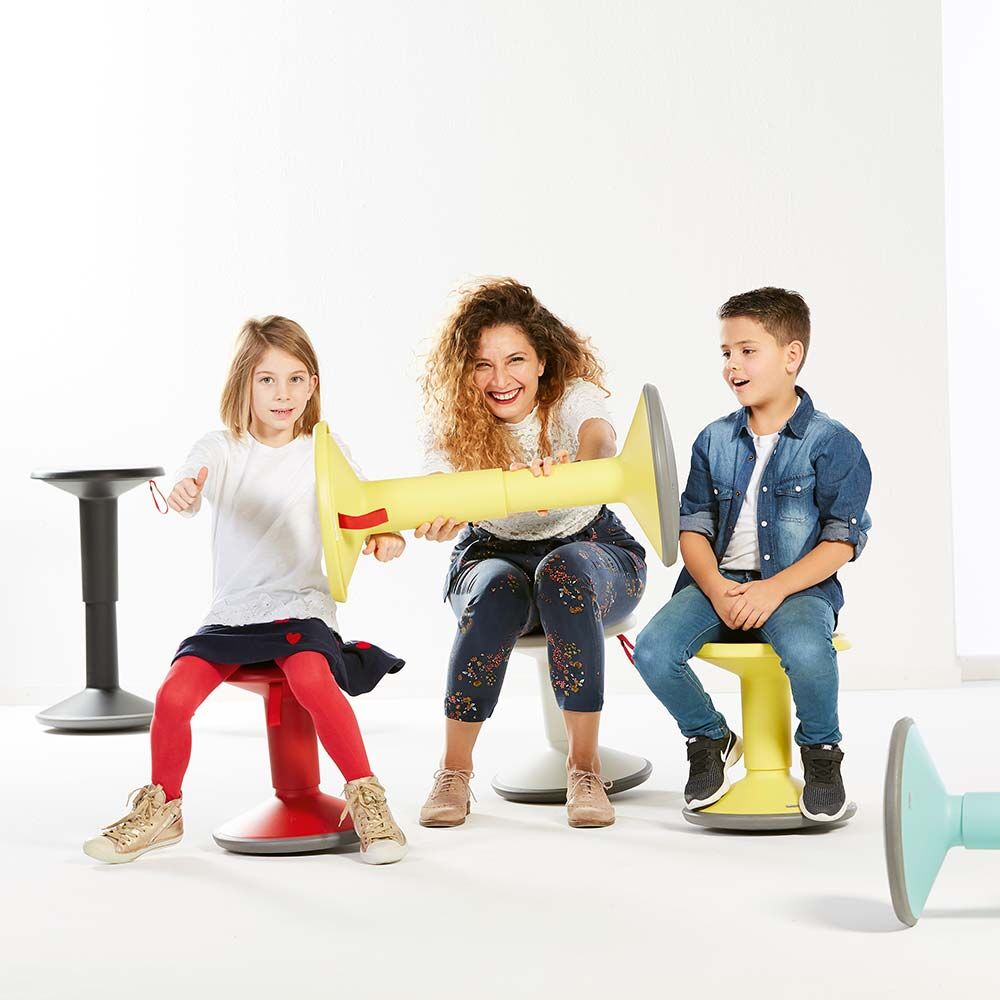 Kinder und eine Frau sitzen auf bunten, ergonomischen Hockern und zeigen dynamisches Sitzen im Büro.