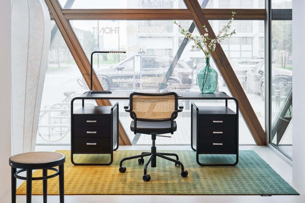 Modernes Büro mit ergonomischem Drehstuhl, Schreibtisch, Schubladenschränken und Glasvase vor großen Fenstern.
