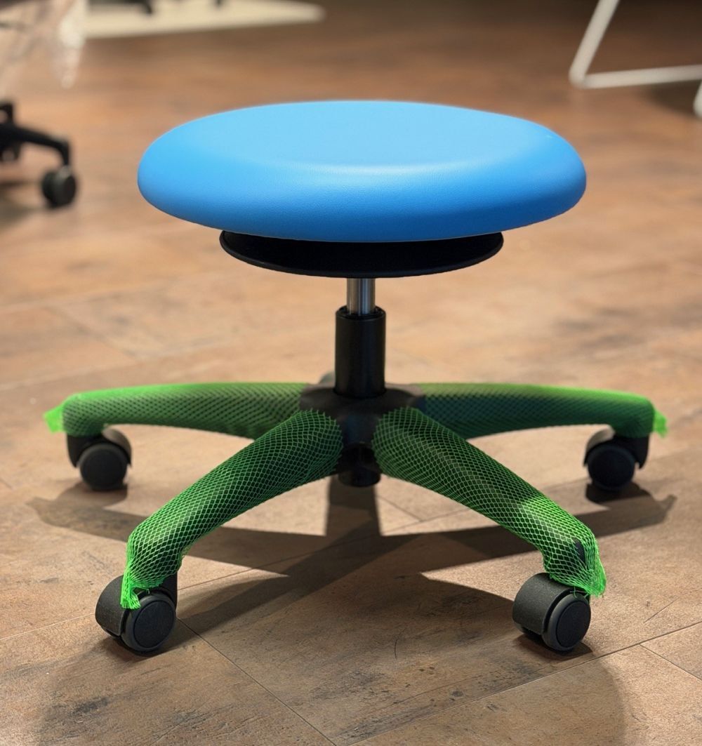 Moderner ergonomischer Hocker mit blauem Sitzpolster und grün umhüllten Rollen auf Holzfußboden.