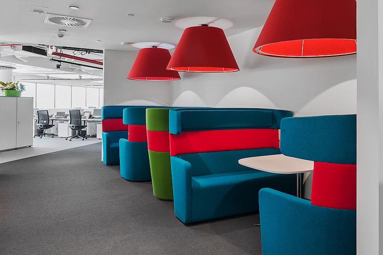 Moderne Büronische mit bunten Polstern, roter Beleuchtung und offenen Arbeitsbereichen im Hintergrund.