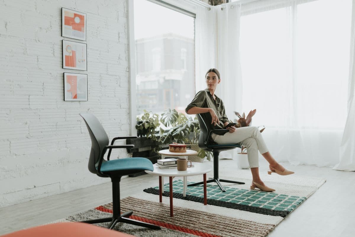 Frau sitzt auf ergonomischem Stuhl in modernem, lichtdurchfluteten Büro mit Pflanzen und künstlerischen Akzenten.