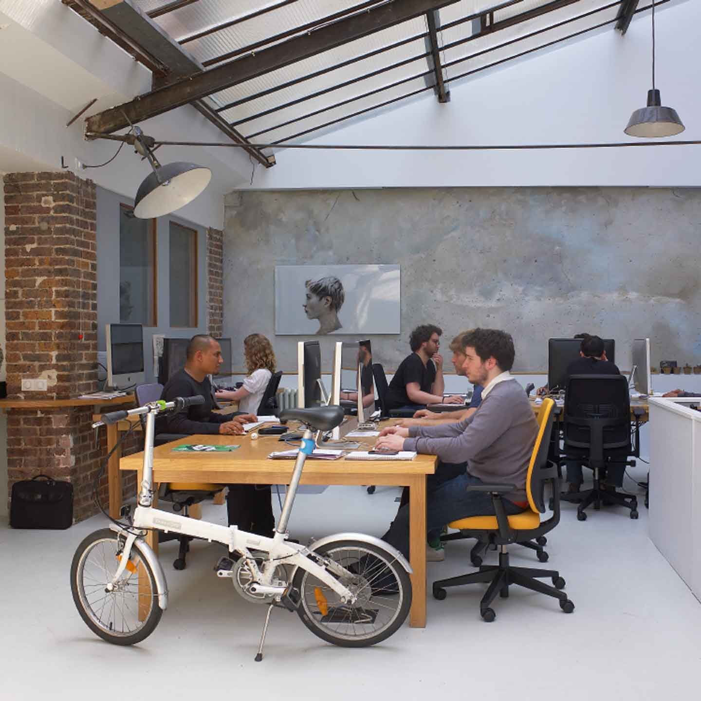 Menschen arbeiten in einem modernen Büro mit ergonomischen Stühlen, unter einem teilweise verglasten Dach.