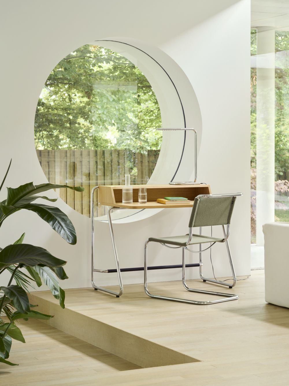 Ergonomischer Schreibtisch mit Stuhl vor rundem Fenster, umgeben von Pflanzen und hellem Holzfußboden.