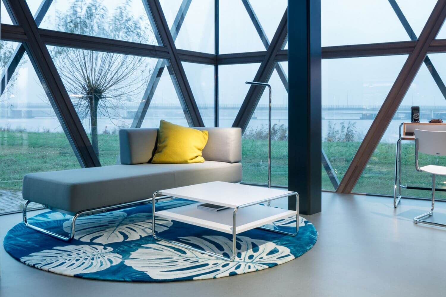 Modernes Wohnzimmer mit grauem Sofa, gelbem Kissen und blauem Teppich vor großen Fenstern mit Naturblick.