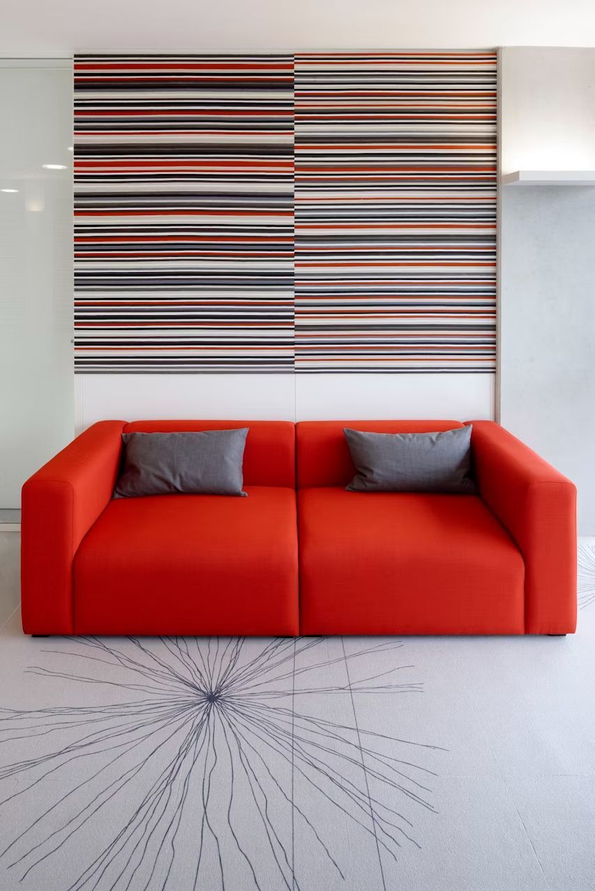 Rotes Sofa mit grauen Kissen vor einer gestreiften Wand in einem modernen Raumdesign.