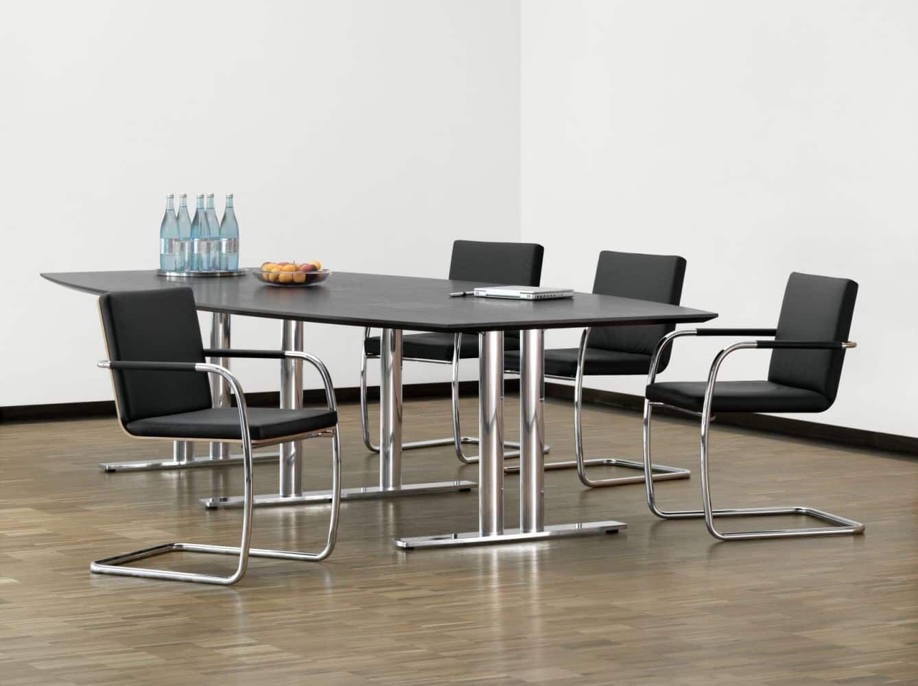 Moderner Konferenztisch mit schwarzen Freischwingerstühlen, Wasserflaschen und Obstdose auf hellem Holzboden.