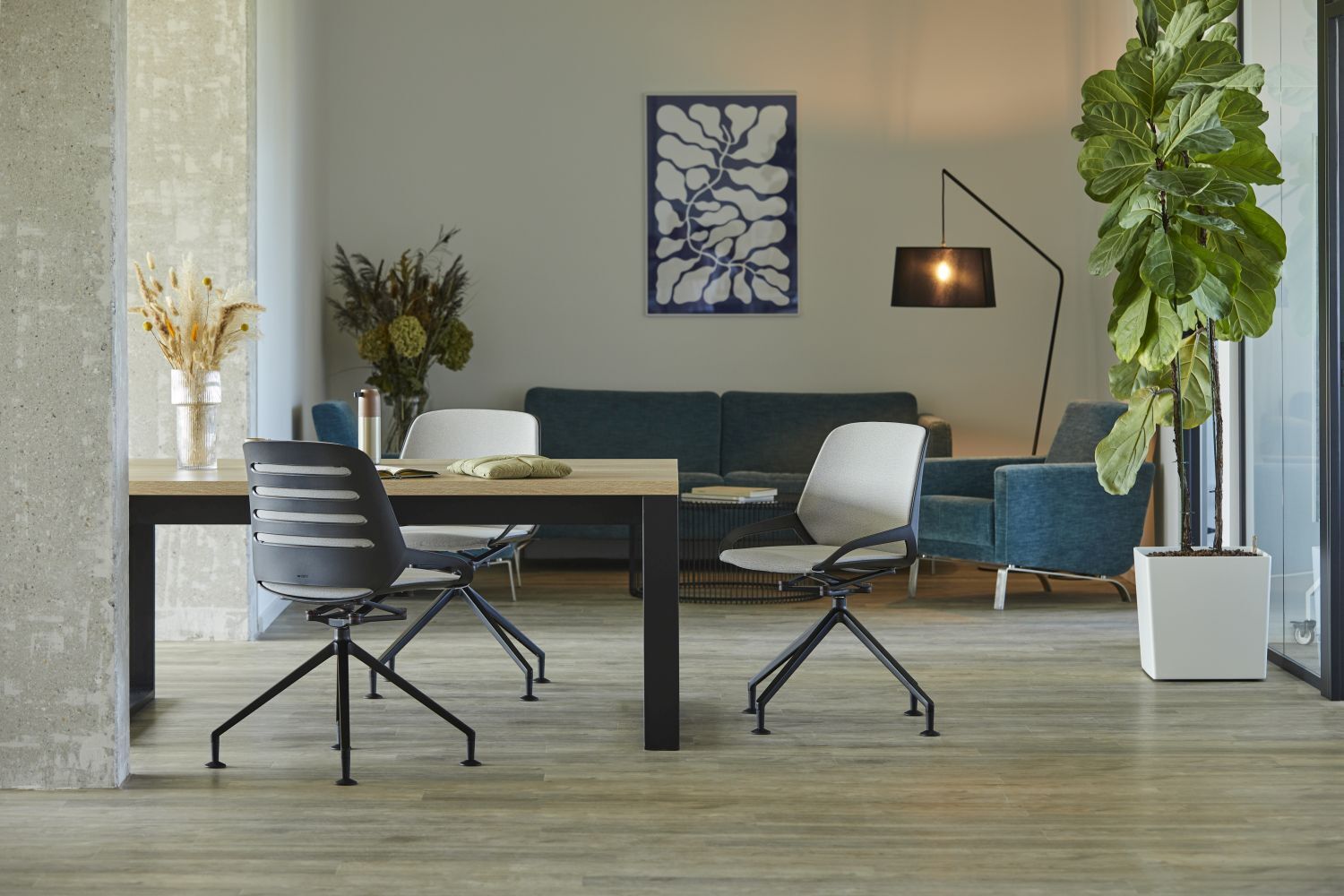 Moderner Büroraum mit ergonomischen Stühlen, Holzschreibtisch, grüner Pflanze und blauer Couch im Hintergrund.