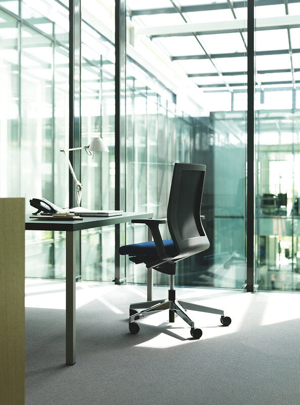 Ergonomischer Bürostuhl am Schreibtisch in einem modernen, lichtdurchfluteten Büro mit großen Glasfenstern.