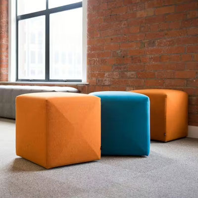 Drei ergonomische Sitzwürfel, zwei orange und ein blau, in einem modernen Büro vor einer Backsteinwand.