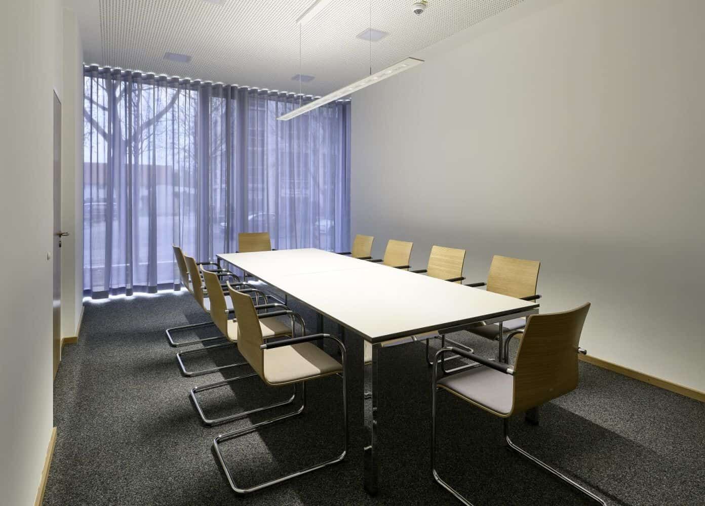 Moderner Besprechungsraum mit hellem Tisch und ergonomischen Stühlen, durchflutet von natürlichem Licht durch große Fenster.