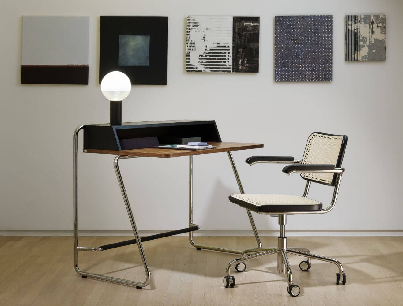 Moderner Schreibtisch mit Lampe und ergonomischem Bürostuhl vor einer Wand mit Kunstwerken.