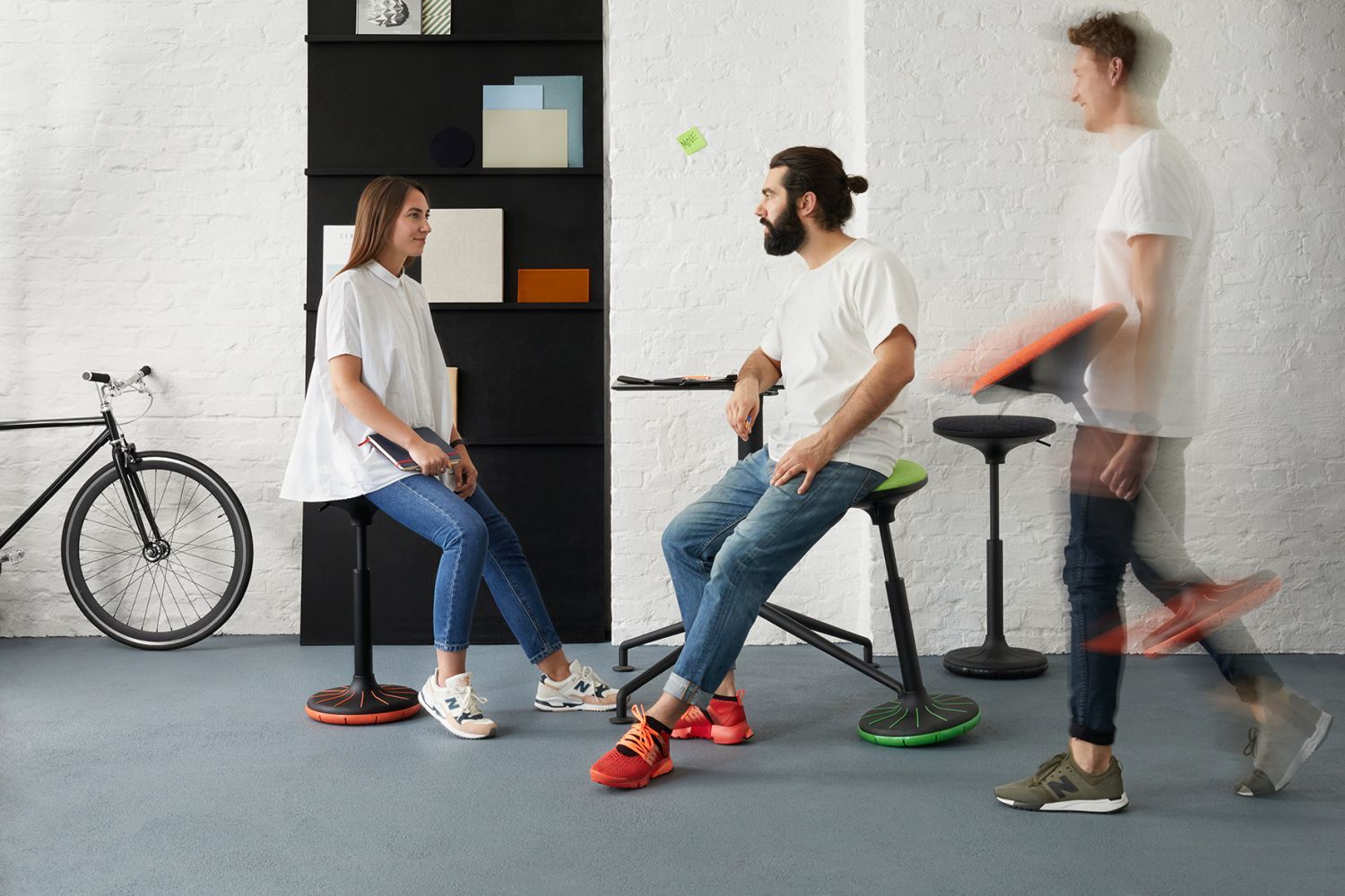 Zwei Personen sitzen auf ergonomischen Hockern, während eine dritte Person mit einem Hocker vorbeigeht.