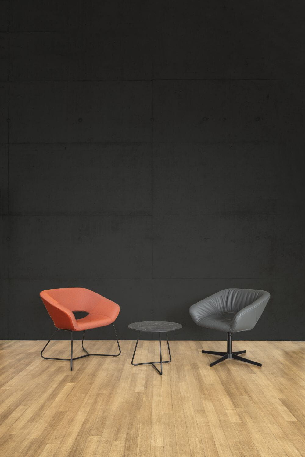 Zwei ergonomische Stühle, einer orange und einer grau, mit einem runden Tisch auf Holzboden vor schwarzer Wand.