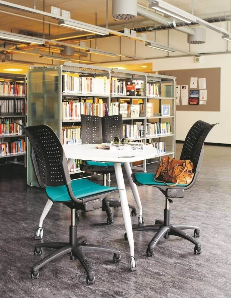 Moderne ergonomische Bürostühle um einen Tisch in einer Bibliothek, mit Regalen im Hintergrund.