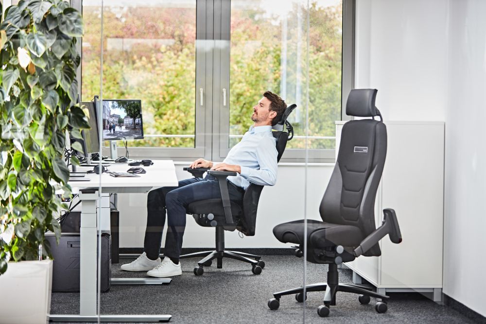Mann sitzt entspannt auf ergonomischem Bürostuhl an einem modernen Schreibtisch vor Fenstern mit Naturblick.