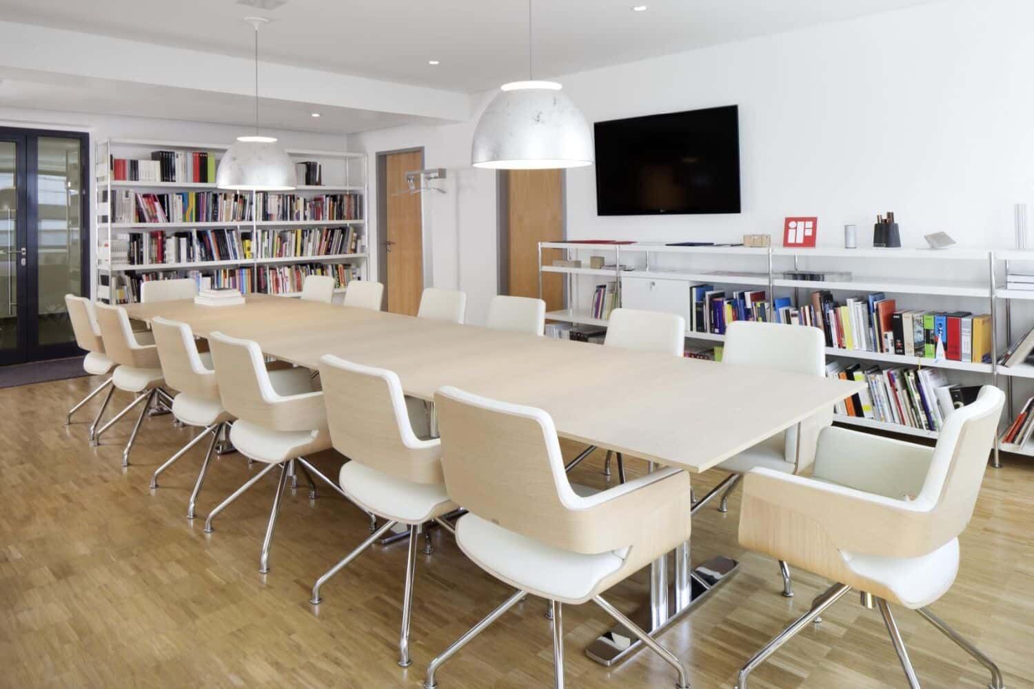 Moderner Konferenzraum mit großem Tisch, ergonomischen Stühlen, Bücherregalen und Wandbildschirm.