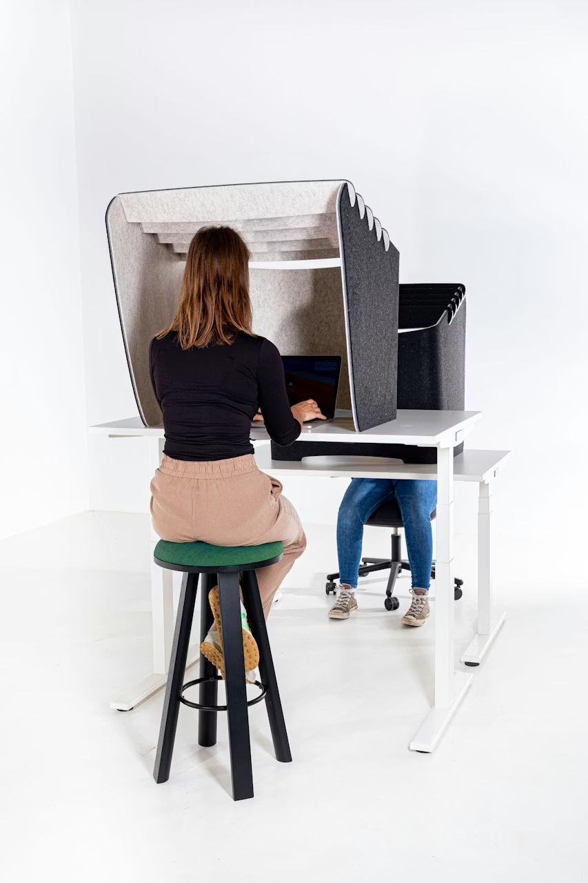 Zwei Personen arbeiten an höhenverstellbaren Schreibtischen mit akustischen Trennwänden, eine auf einem Hocker, die andere auf einem Bürostuhl.