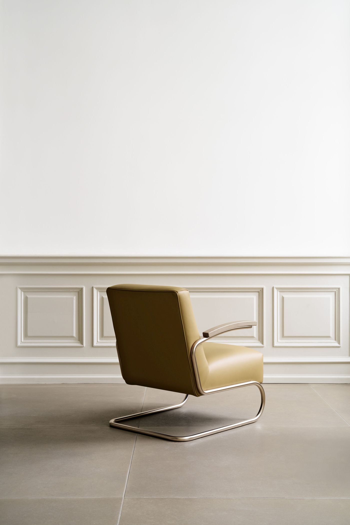 Beiger, ergonomischer Sessel mit Metallgestell vor einer eleganten, weißen Wand mit klassischer Verkleidung.