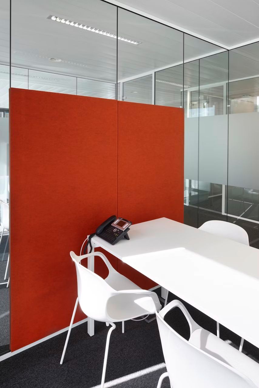 Moderner Büroarbeitsplatz mit weißem Tisch, drei Stühlen und orangener Trennwand in einem Glaskabinenraum.