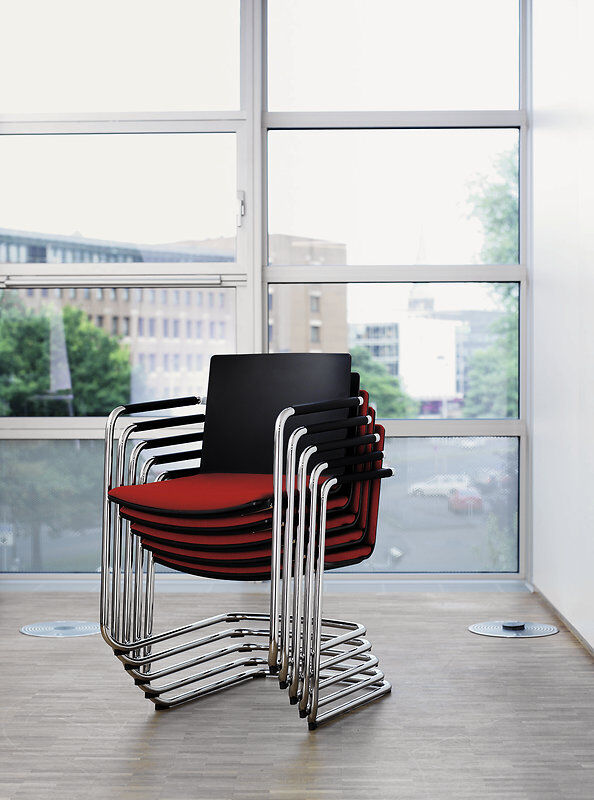 Gestapelte ergonomische Stühle mit rotem Sitzpolster und verchromtem Gestell, in einem modernen Büro mit Fenstern.