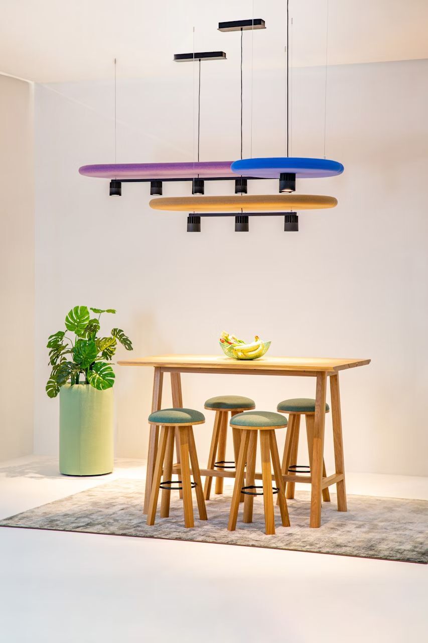 Heller Raum mit Holztisch, vier Hockern, einer Pflanze und bunten, modernen Pendelleuchten an der Decke.