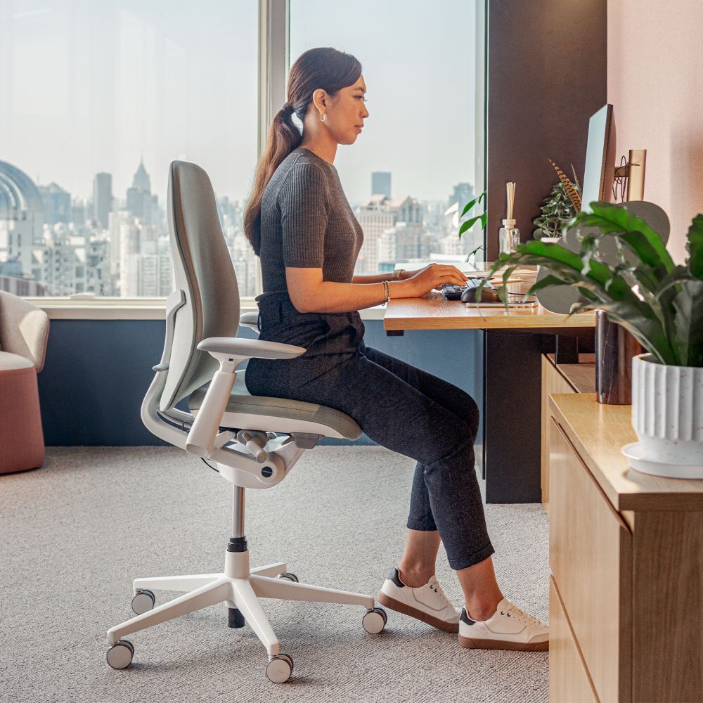 Frau sitzt ergonomisch an einem Schreibtisch in einem modernen Büro mit Stadtblick, gesunder Sitzhaltung.